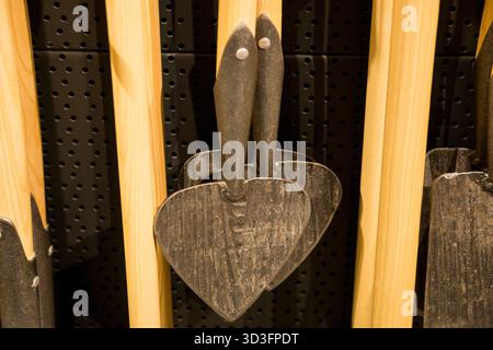 Primo piano di pale da giardino in metallo a forma di cuore appese su manici in legno su sfondo nero perforato in un negozio di ferramenta. Foto Stock