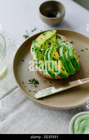 Avocado tagliato a fette su pane tostato integrale con erbe e semi freschi, servito su un piatto di ceramica beige con un coltello, e sistemato su un panno di lino con un recipiente Foto Stock