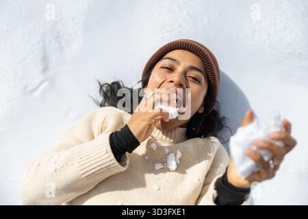 Una donna gioiosa si stende su una coperta di neve nei Pirenei, portandole giocosamente la neve in bocca, vestita calorosamente con un maglione in maglia e un berretto, avvolgente Foto Stock