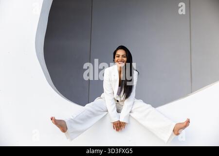 Una donna d'affari sicura di sé in un elegante abito bianco si trova graziosamente su un suggestivo sfondo urbano moderno, incarnando professionalità e stile in un Foto Stock