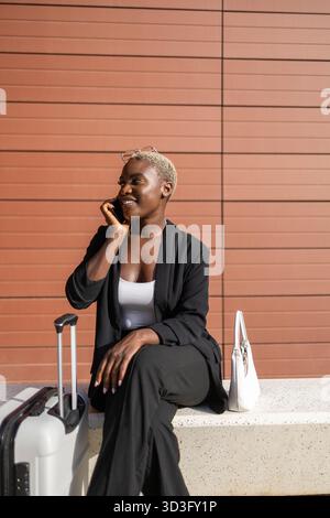 Una donna d'affari professionista siede con fiducia su una panchina, impegnata in una telefonata. Vestita con un vestito nero con un top bianco, ha una valigia e una h Foto Stock