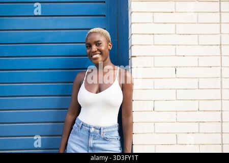 Una donna d'affari e CEO sicura di sé si trova casualmente contro un vivace muro blu, trasudando professionalità e disponibilità in un semplice top bianco e un j Foto Stock
