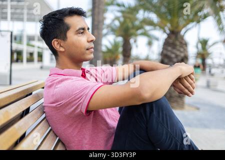 Un uomo ispanico attento siede pensiamente su una panchina di legno in un ambiente esterno. Vestito casualmente con una camicia rosa e jeans blu, sembra essere en Foto Stock