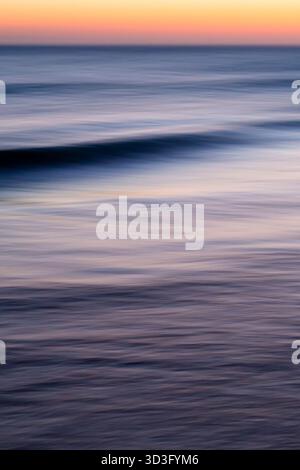 Le onde dolci si infrangono su un mare tranquillo mentre il sole tramonta, generando una calda tonalità dorata sull'acqua. L'immagine cattura la bellezza serena e pacifica Foto Stock