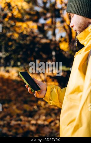 Un uomo con un impermeabile giallo è immerso in una vivace vegetazione autunnale, concentrata sul suo smartphone. La scena cattura l'essenza della tecnologia che si fonde con Foto Stock