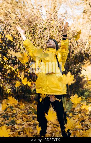 Un uomo allegro che indossa un impermeabile giallo brillante lancia con gioia le foglie autunnali in aria, circondato dai colori vivaci del fogliame autunnale in un parco Foto Stock