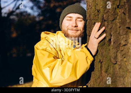 Un uomo con un impermeabile giallo e un cappello a maglia abbraccia un albero, gli occhi chiusi, assaporando la bellezza tranquilla di una giornata autunnale nel parco. Filtri solari t Foto Stock