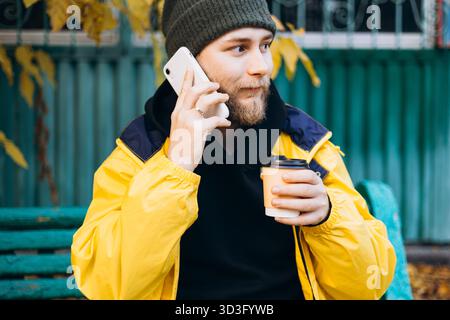 Un uomo che indossa un impermeabile giallo si siede su una panchina, gustando una bevanda calda durante una giornata autunnale. Il suo abbigliamento casual si abbina perfettamente al vibrante fogliame Foto Stock