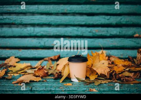 Una tazza da caffè usa e getta si appoggia tra le colorate foglie autunnali su una panca in legno blu rustico, catturando l'essenza dell'autunno e un momento accogliente e tranquillo Foto Stock