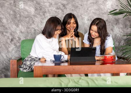 Tre adolescenti asiatici sono impegnati e sorridenti mentre si riuniscono intorno a un tablet in un caffè indonesiano alla moda, godendosi l'un l'altro in un ambiente urbano Foto Stock