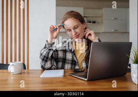 Una donna premurosa che indossa gli occhiali si siede a un tavolo di casa utilizzando un laptop e uno smartphone, multitasking con la tecnologia. Sembra concentrata sulla bacchetta remota Foto Stock
