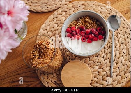 Un'accogliente ciotola di muesli fatti in casa condita con vivaci bacche rosse e cremoso yogurt si trova su un tappetino intrecciato, che offre una deliziosa e salutare colazione Foto Stock