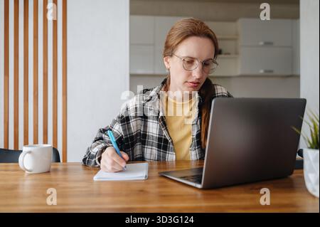 La donna che indossa gli occhiali utilizza un notebook e scrive su un notebook a un tavolo di casa, mostrando il multitasking con la tecnologia. Ideale per rappresentare remo moderno Foto Stock