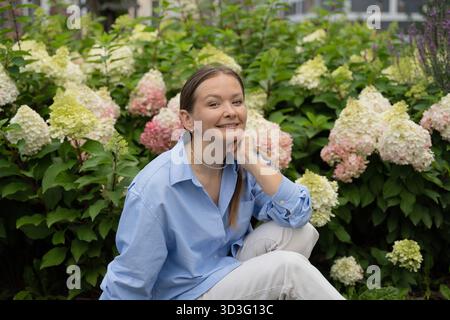 Una giovane donna che indossa una camicia blu si posa felicemente in mezzo a ortensie vibranti in un moderno giardino cittadino, catturando un momento di serenità e bellezza nella città Foto Stock
