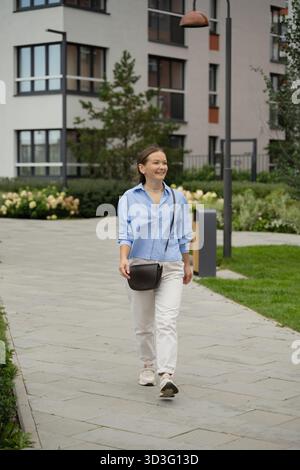 Una giovane donna cammina con sicurezza attraverso un paesaggio urbano moderno, indossando una camicia casual blu e pantaloni bianchi. Lo sfondo presenta un arco contemporaneo Foto Stock