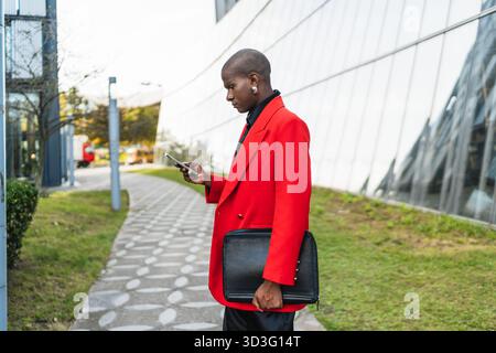 Un giovane uomo LGBTQ in un vivace blazer rosso si erge all'aperto, vicino a un edificio moderno, guardando il suo telefono, tenendo un portafoglio nero, simboleggiando la confidenc Foto Stock