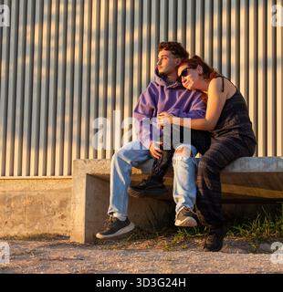 Una coppia LGBT siede su una panchina, avvolta tra le braccia dell'altra, bagnata dalla luce dorata dell'ora. Le loro espressioni pacifiche riflettono un momento condiviso Foto Stock