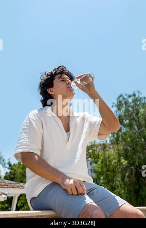 Un giovane viene catturato da un angolo basso bevendo acqua sotto un cielo azzurro. È seduto all'aperto, indossa abiti casual e si gode una rinfrescante Foto Stock
