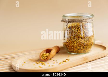 Un vaso di vetro pieno di polline naturale d'api si trova su un vassoio di legno con una piccola paletta di legno, splendidamente sistemata su un morbido sfondo beige, alto Foto Stock