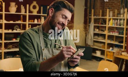 Felice uomo caucasico ragazzo maschio proprietario di una piccola impresa imprenditore potter artigiano multitasking scrittura note scrivere piano parlare telefono cellulare chiamata chat Foto Stock