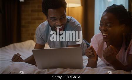 Felice eccitato afroamericano ragazza fidanzato uomo sdraiato sul letto famiglia coppia guardare tablet digitale scioccato sport partita partita vittoria Foto Stock