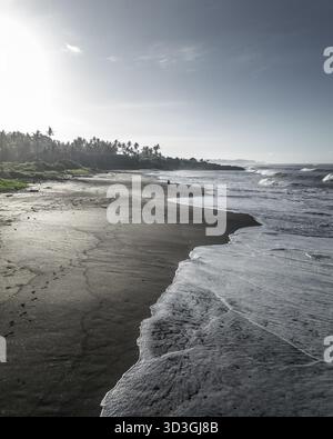 Vista aerea della spiaggia di sabbia nera con onde bianche schiumose che contrastano con la sabbia vulcanica scura sotto il sole luminoso, Bali, Indonesia. Foto Stock