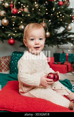 Bambino che festeggia il Natale vicino all'albero festivo. Bimbo sorridente in maglia di panna con ornamenti rossi seduto davanti a un Cristo decorato Foto Stock