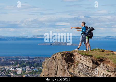 Due avventurieri sono appollaiati su una scogliera, sulla cima del monte Arthurs Seat, Edinburg, che punta verso una vasta scena urbana e il vasto Water bey Foto Stock