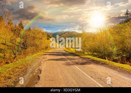 strada attraverso il paesaggio montano in autunno al tramonto. foresta dai colori autunnali sotto il cielo blu e l'arcobaleno alla luce della sera. picco innevato in località dista Foto Stock