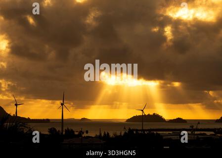 I suggestivi raggi dorati del sole penetrano attraverso nuvole buie e pesanti su un paesaggio marino caratterizzato da turbine eoliche sagomate e piccole isole al tramonto. Foto Stock