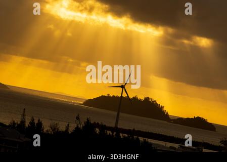 I suggestivi raggi dorati del sole penetrano attraverso nuvole buie e pesanti su un paesaggio marino caratterizzato da turbine eoliche sagomate e piccole isole al tramonto. Foto Stock