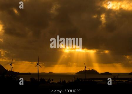 I suggestivi raggi dorati del sole penetrano attraverso nuvole buie e pesanti su un paesaggio marino caratterizzato da turbine eoliche sagomate e piccole isole al tramonto. Foto Stock