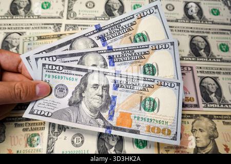 In questa illustrazione fotografica, una mano tiene in mano diverse nuove banconote da 100 dollari, con uno sfondo di varie altre banconote statunitensi. Foto Stock