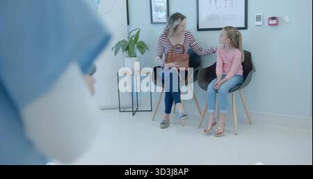 Madre seduta in maglione a righe e figlia in cima rosa nella sala d'attesa della clinica, con borsetta Foto Stock