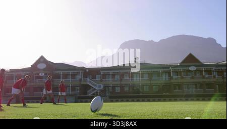 Posizionamento di cinque giocatori di rugby che indossano maglie rosse per preparare il calcio sul campo scolastico, con palla sul tee Foto Stock