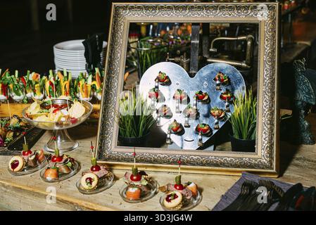 Cibo, elegante tavolo a buffet con stuzzichini assortiti, tazze di verdure e un creativo display a cuore metallico incorniciato in una cornice decorativa dorata Foto Stock