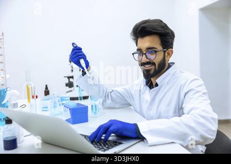 Uomo scienziato in camice da laboratorio con contagocce in vetro con reagenti, prendere appunti in computer portatile, sperimentare campioni medici in laboratorio Foto Stock