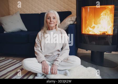 Donna anziana con capelli grigi seduta sul divano in soggiorno con camino Foto Stock