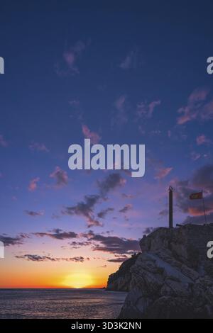 Statua obelisco e bandiera montenegrina che fluttua nel vento nella fortezza sulla cima della scogliera a Petrovac costa al tramonto, Montenegro Foto Stock
