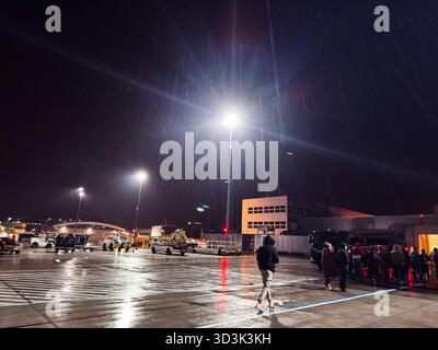 Balice, Polonia - 4 ottobre 2025: Scena notturna all'aeroporto di Cracovia con viaggiatori in fila, luci accese, pioggia e asfalto bagnato. Foto Stock