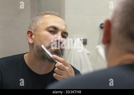 Bell'uomo di mezza età che si lava la barba la mattina in bagno guardando nello specchio. Concetto di cura del viso Foto Stock