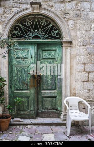 Sedia in plastica bianca di fronte bloccato con lucchetti e catene verde vecchio danneggiato porte in legno in uno degli edifici di Kotor, Montenegro Foto Stock