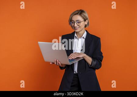 Donna d'affari sicura che indossa un abito nero di stile ufficiale che lavora su un laptop in ufficio isolato su sfondo arancione Foto Stock