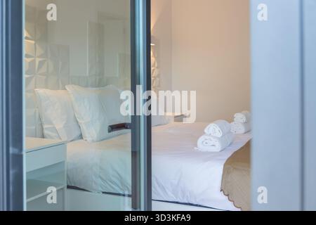 Vista dall'esterno di una camera d'albergo o di una pensione con un letto ben fatto, asciugamani piegati e una coperta beige, che rappresenta l'ospitalità Foto Stock