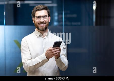 Ritratto di un giovane specialista di uomini sorridenti che si trova in un ufficio di lavoro, in piedi, tenendo un telefono e guardando la fotocamera. Foto Stock