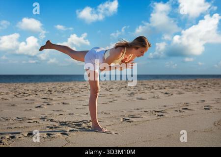 Una donna regala graziosamente il Virabhadrasana Warrior 3 posa sulla spiaggia, bilanciandosi su una gamba. Il sole tramonta sullo sfondo, proiettando colori caldi Foto Stock