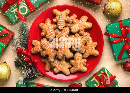 Vista dall'alto del piatto rosso con biscotti dell'uomo di pan di zenzero natalizio e ornamenti natalizi su una tovaglia color oro glitter. Concetto di dessert di Natale Foto Stock