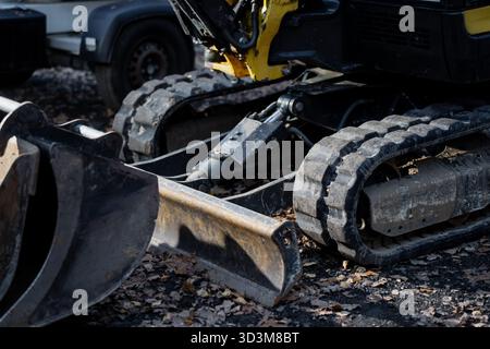 Escavatore compatto con cingoli in gomma e lama di livellamento in acciaio parcheggiata in cantiere all'aperto ricoperta da foglie asciutte che mostrano i dettagli dei macchinari da costruzione Foto Stock