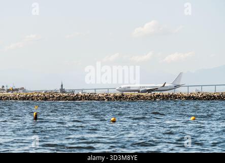 Aereo passeggeri rullante vicino alla pista vicino al mare con montagne sullo sfondo, vista dall'acqua con boe e cielo limpido, concetto di viaggio, aeroporto i. Foto Stock