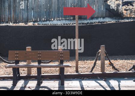 Mockup con segnaletica a freccia rossa vuota su un'asta di legno accanto alla panca vuota nel soleggiato parco all'aperto, modello perfetto per la direzione, la navigazione o la segnaletica Foto Stock